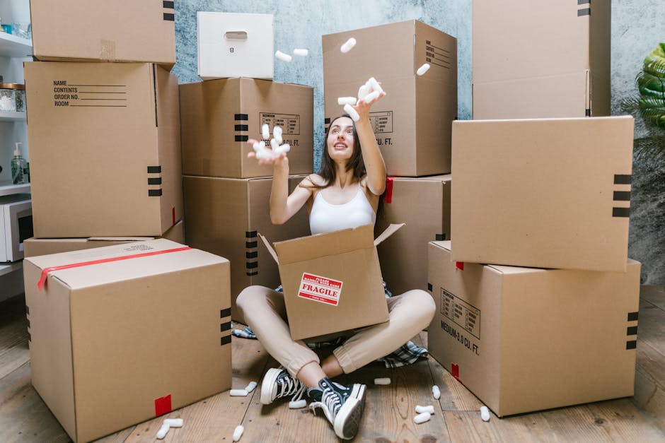 A young woman sitting on a wooden floor surrounded by multiple cardboard moving boxes of various sizes, some labeled with handling instructions such as 'fragile', inside an interior space with light b